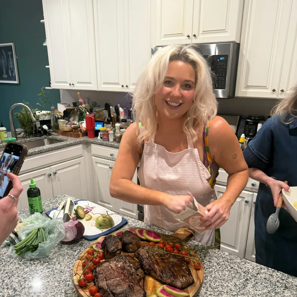 Kaylee in an apron behind a board of sliced steak with watermelon radish and blistered tomatoes, mid-service in a home kitchen