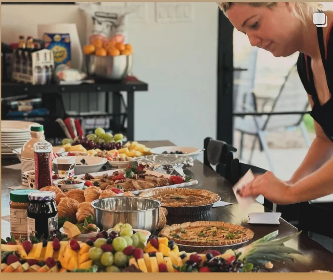 Kaylee in a black apron leaning over a long table of brunch — herb-topped tarts, croissants, cheese and charcuterie, and a platter of grapes, pineapple and berries — finishing a dish while the spread is still going up