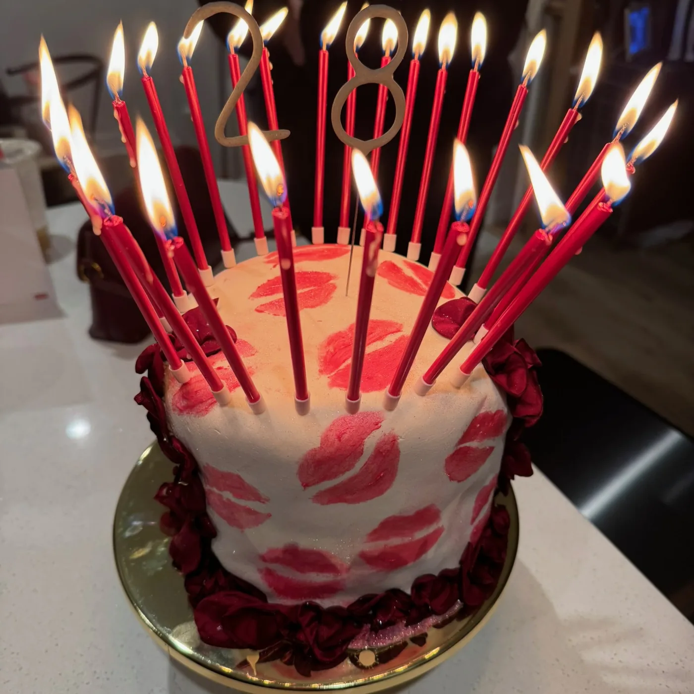 A white birthday cake painted with red lipstick kisses, ringed with dark red rose petals and lit with a crown of tall red candles.