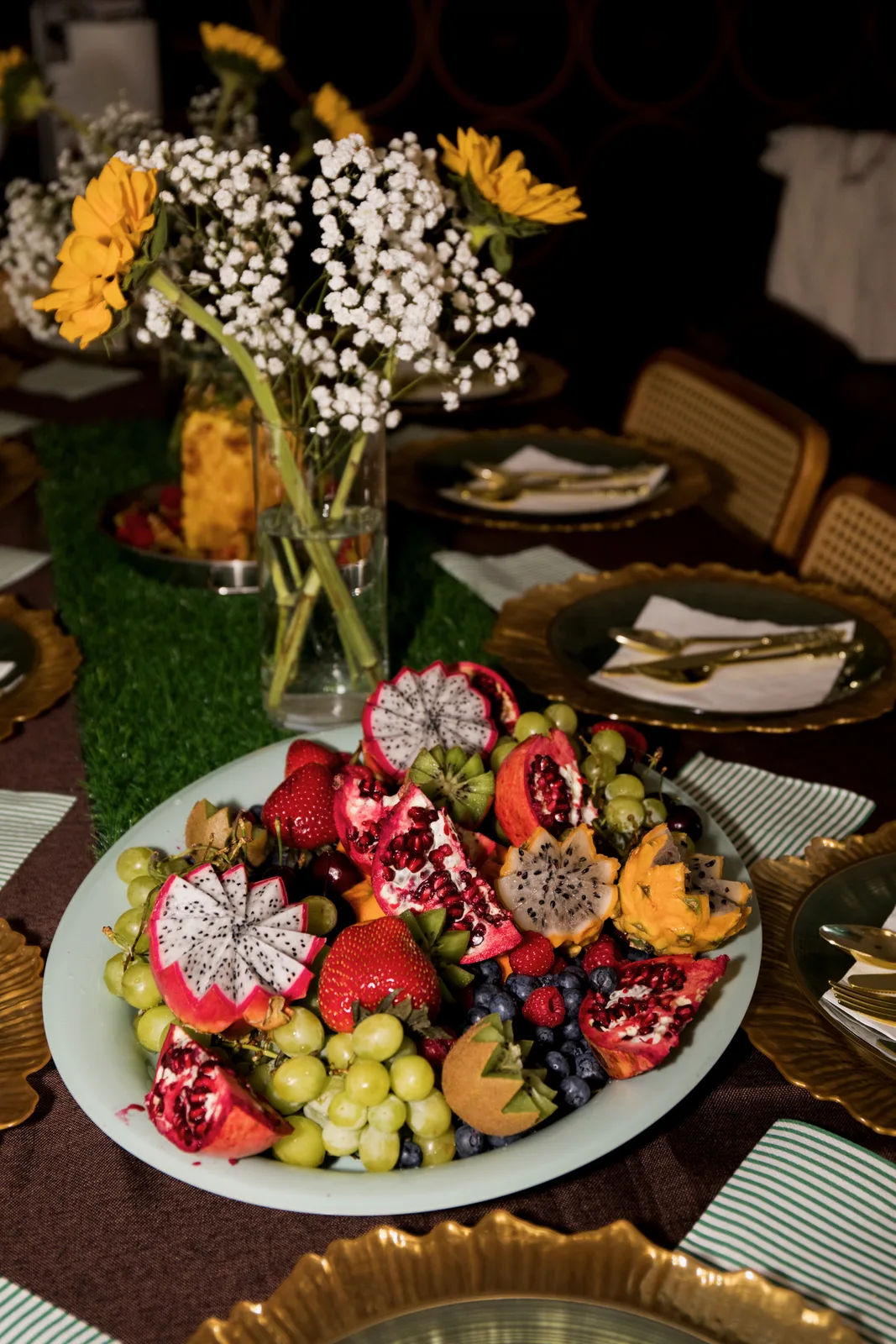A platter of cut fruit — dragon fruit, pomegranate, strawberries and grapes — on a dressed table with gold chargers, striped napkins and sunflowers, lit low.