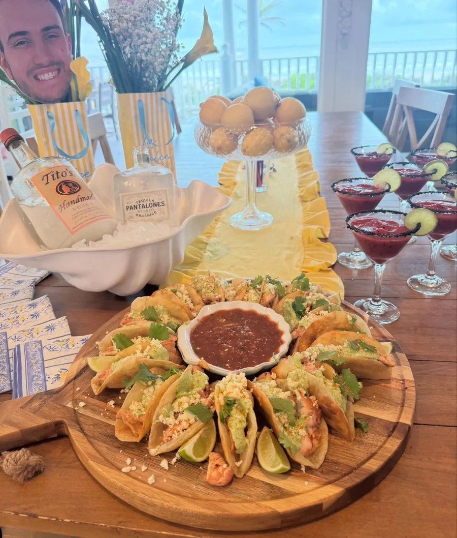 A round board of shrimp tacos with salsa, a row of salt-rimmed margaritas and tequila on ice, on a table overlooking the water.