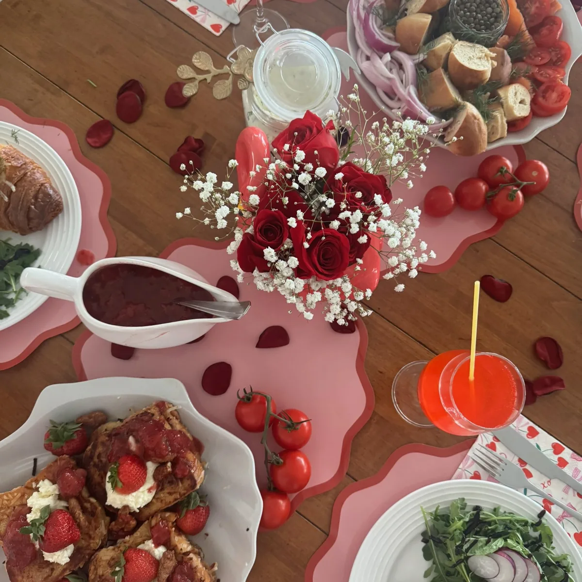 A table laid for two in reds and pinks — red roses and baby's breath, brioche with strawberries and ricotta, a jug of sauce and a bright cocktail.